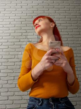 Young red-haired girl is reading something thoughtfully on her smartphone against a gray brick wall