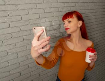 Young red-haired girl with a cup of coffee makes selfie with her smartphone on a background of gray brick wall