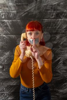 Conceptual picture. Young red-haired girl tries to talk on the phone with sealed with adhesive tape with her lips so that she keeps silence