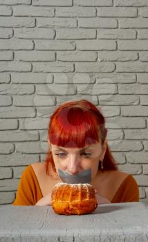 Conceptual image of a young red-haired girl on a diet sitting with her mouth taped with silver tape before appetizing pastries