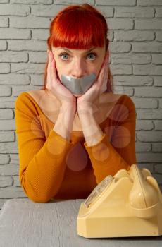 Conceptual picture. Young red-haired girl sitting near an old dial-up phone with adhesive tape lips to keep silence