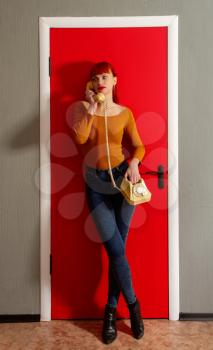 Pretty woman talking on an old dial telephone with a twisted wire on the background of a bright red door