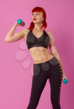 young red-haired girl in sportswear engaged with dumbbells on a pink background