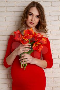 Beautiful young girl in the sixth month of pregnancy in a bright red dress with a bouquet of tulips on the background of a brick wall.