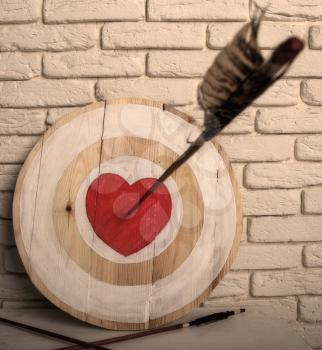 Handmade rough wooden target with a center in the form of a red heart and an arrow from a bow that hit the center and two arrows missed.