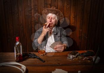 elderly gray-haired cowboy enjoying playing cards sipping whiskey in a dark saloon at a wooden table