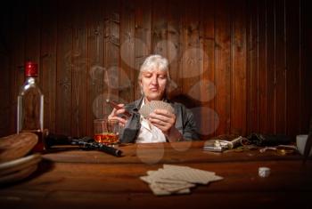 elderly gray-haired cowboy enjoying playing cards sipping whiskey in a dark saloon at a wooden table