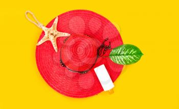 Elegant female red beach hat lies on a yellow background next to a starfish with sunblock and a green leaf