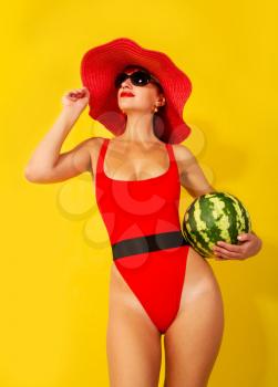 young girl in a bright red swimsuit and a wide-brimmed red hat eats a watermelon on a yellow background