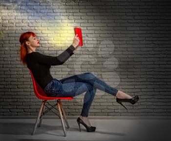 young girl in casual clothes reads an interesting or magical book leaning back in a chair against a dark brick background