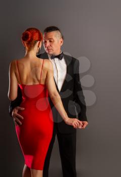 A man in a strict dark suit and a beautiful girl in a bright red dress are dancing a slow sensual dance on a dark background.