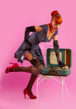 Pin-up model girl in retro polka-dot dress listens to an old gramophone on a pink background