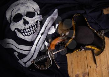 Classic pirate black felt captain's cocked hat with a pistol and a sword lying on a wooden floor next to the Jolly Roger flag
