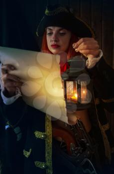Young Attractive armed girl pirate captain examines a map in the light of a lantern against the backdrop of the flag