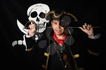 a pirate captain in a camisole and a cocked hat stands with a sword in his hands against the background of a black flag jolly roger.