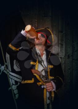 Adult pirate captain in a traditional costume and with weapons drinks rum from a clay bottle against the background of a jolly roger