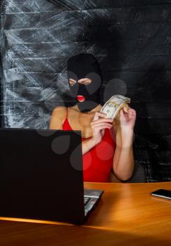 young girl in a sexy red dress and a balaclava mask sits at a laptop and makes an honest profit on the Internet