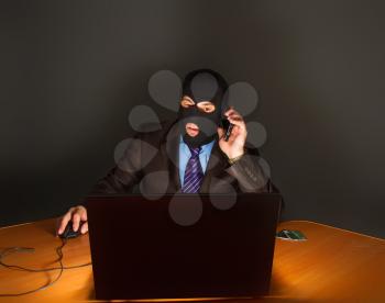 representative businessman in a suit and tie, but in a mask with a balaclava covering his face, is engaged in theft through the Internet from a laptop