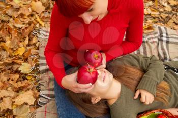 young mother and little daughters in bright sweaters on a picnic in nature indulge in apples