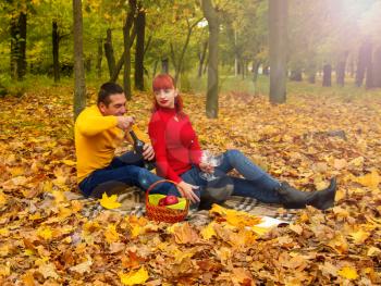 husband and wife in bright clothes for relaxing settled down on a picnic among the trees and fallen leaves with a bottle of wine and glasses.