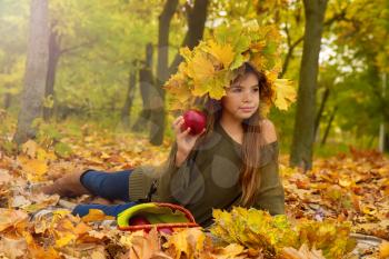 Little girl on a picnic in autumn park eats a ripe red apple sitting on fallen foliage in a headdress from maple leaves