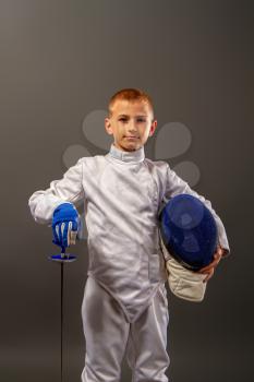 little boy fencer armed with a rapier in white sports equipment and with a protective helmet on a dark background