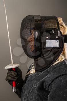 young girl a fencer in black coaching equipment and a black mask on her face stands holding up a rapier preparing for battle on a dark background.