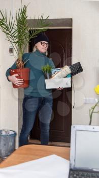 Sad adult woman fired from work is standing near the office door with a box of belongings and a beloved palm tree.