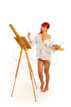 A young red-haired girl in a paint-soaked shirt sits in front of an easel with an empty canvas with a palette and brushes