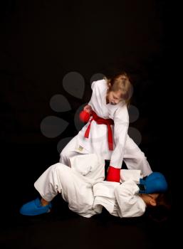 Two little karateka in white kimonos, one in red, the other in blue competition equipment fight against a dark background