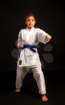 Little girl in a sports kimono and a blue belt performs exercises in kata on a dark background