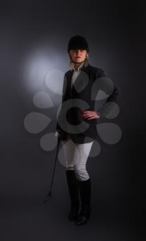 woman rider in a hard protective helmet and a riding suit with a whip in hand posing on a gray background