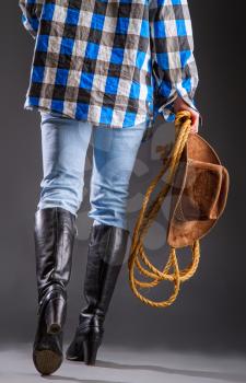Legs in jeans and boots of a cowboy girl in a checkered shirt with a lasso and a wide-brimmed hat on a dark background