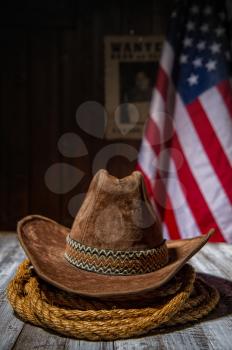 Classic cowboy hat and lasso lie on a wooden table against the background of the US flag and the poster for the search for the criminal in the sheriff s office.