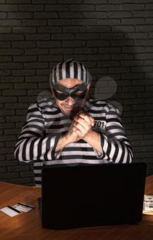 funny petty crook in exaggerated prisoner's clothes and a black mask with a silly face steals money on the Internet from gullible users through a laptop.