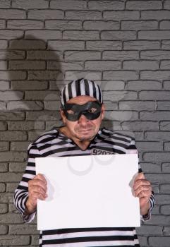 funny petty crook in exaggerated clothes of a prisoner and a black mask with a silly face holds an empty sign in the hands of a brick wall.