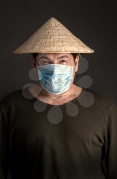 Chinese adult man in traditional Asian conical straw hat stands in virus-protected medical mask on a dark background