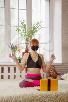 Young girl in medical mask practices yoga at home during quarantine via the Internet.