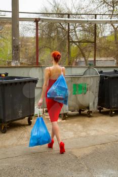 Stylish young girl in an evening dress and in red high-heeled shoes with bags of garbage found the only opportunity to walk during the virus.