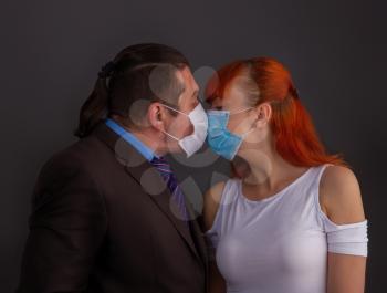 A man in a suit and a girl kiss on a dark background through protective medical masks.