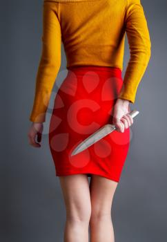 A young girl in a bright skirt and blouse hides a large steel kitchen knife behind a back in her arms on a dark background