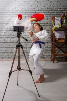A little girl in kimono is engaged in online karate training on a mobile device with an imaginary opponent who is trying to hit through the screen.