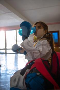 Tired karate girl in a white kimono and blue outfit drinks water from a bottle after a fight in a desert gym