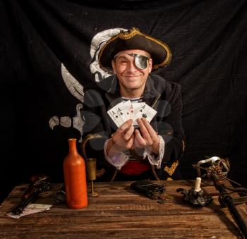 an adult pirate in a cocked hat and a striped vest plays cards and a wooden table against the background of the Jolly Roger flag