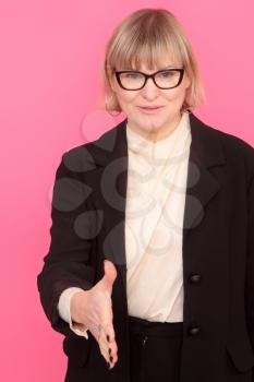 business woman in a formal suit and glasses affably extends her hand to her interlocutor on a pink background