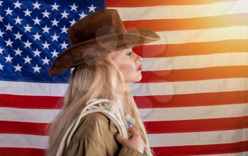 A blonde patriotic lady in a cowboy hat and with a lasso stands against the background of the star-striped flag of the United States of America 