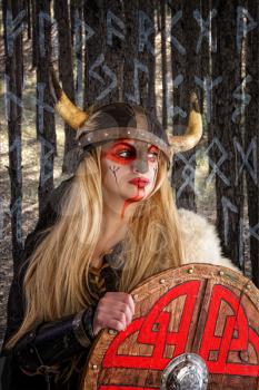 A beautiful blonde viking girl in a horned helmet is pensively standing with a shield on the background of the forest and the background from the Scandinavian alphabet 