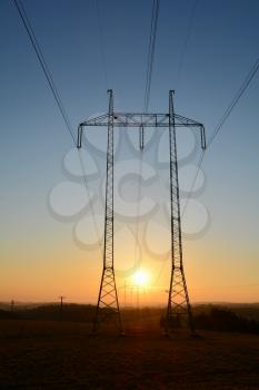 High voltage power lines with big and tall pylons during sunset. The sun sets between two pylons.