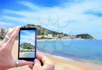 View over the mobile phone display during taking a picture of Costa Brava beach. Holding the mobile phone in hands and taking a photo. Focused on mobile phones screen. 