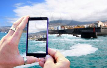 View over the mobile phone display during shooting of Tenerife coast. Holding the mobile phone in hands and taking a photo, focused on mobile phone screen.